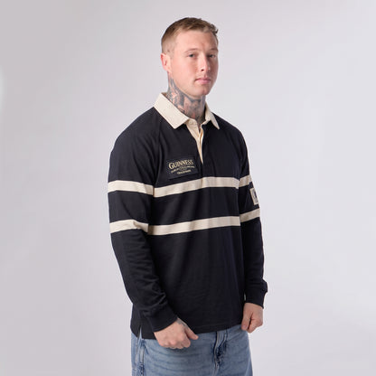 A man with tattoos stands against a plain light background wearing the Guinness Black Traditional Rugby Jersey by Guinness, crafted from 100% cotton in black and cream.