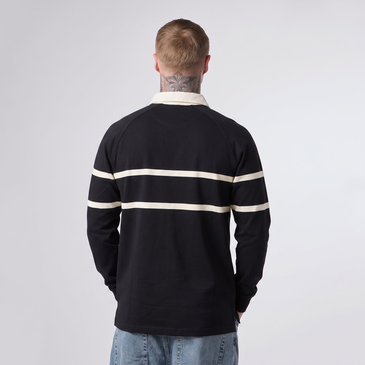 A person with short hair and a neck tattoo is standing with their back to the camera, wearing the Guinness Black Traditional Rugby Jersey by Guinness along with light blue jeans.
