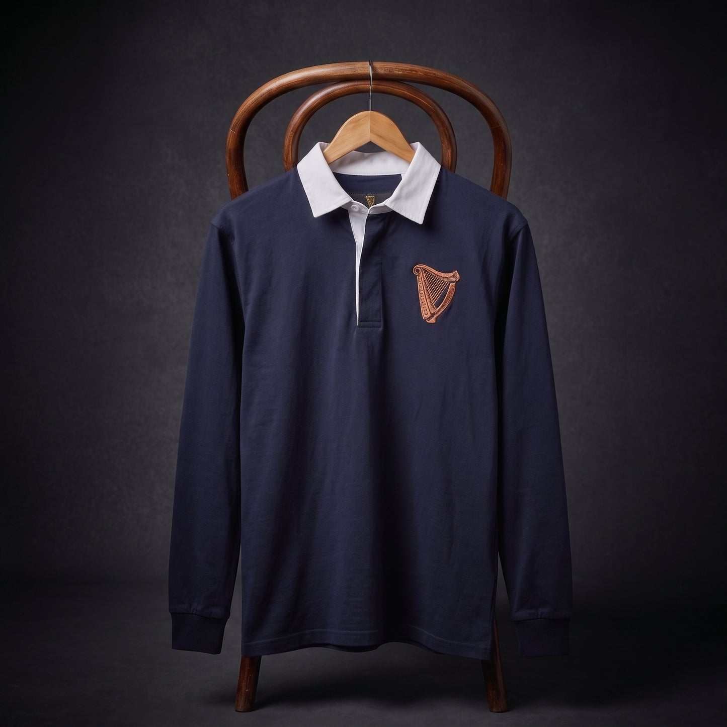The Guinness Navy Traditional Rugby Jersey from the Guinness Webstore US is a long-sleeve polo inspired by classic rugby designs, featuring a crisp white collar and a gold harp emblem on the chest. Displayed on a wooden hanger and chair against a dark background, it captures the timeless style of an authentic Guinness rugby jersey.
