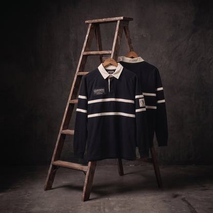 Two Guinness Black Traditional Rugby Jerseys—classic examples of rugby apparel from Guinness—are displayed on hangers, one in front of and one behind a wooden stepladder, set against a dark, textured background.