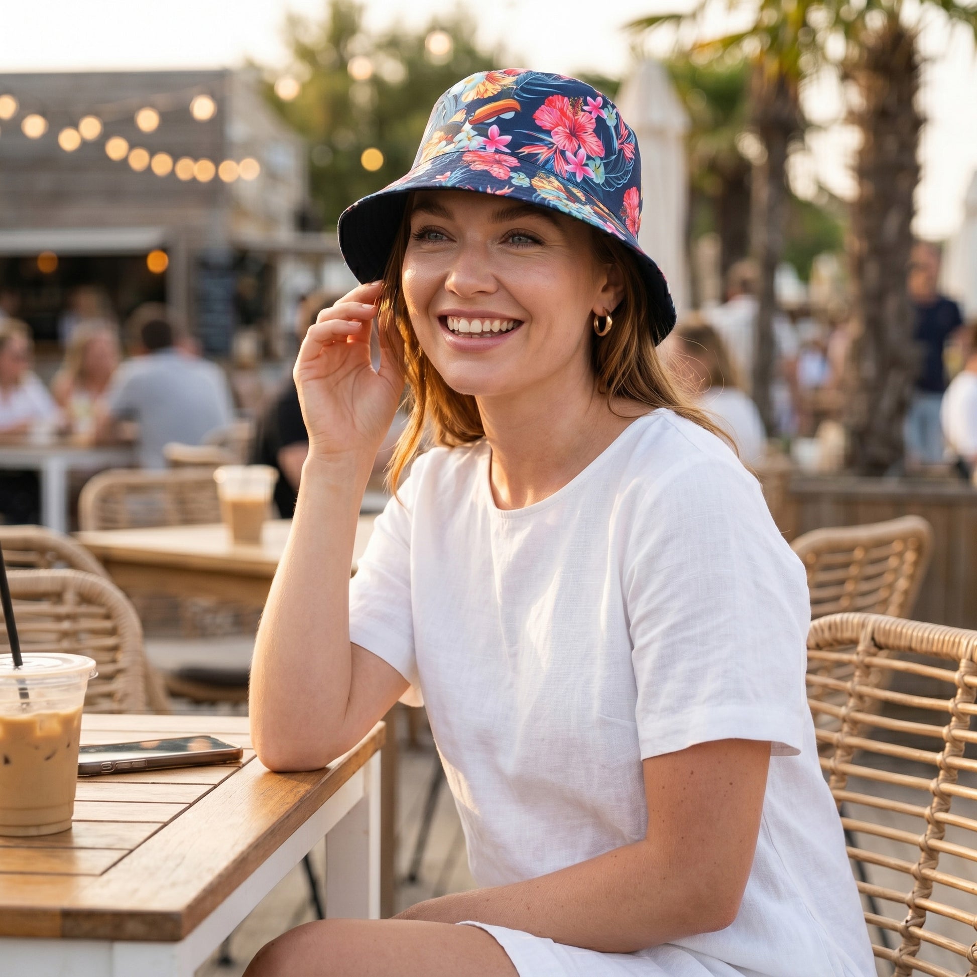 A woman in a white dress and the Guinness Hawaiian Reversible Bucket Hat from Guinness Webstore US sits smiling at an outdoor café table with iced coffee and her phone, while people and palm trees fill the background.