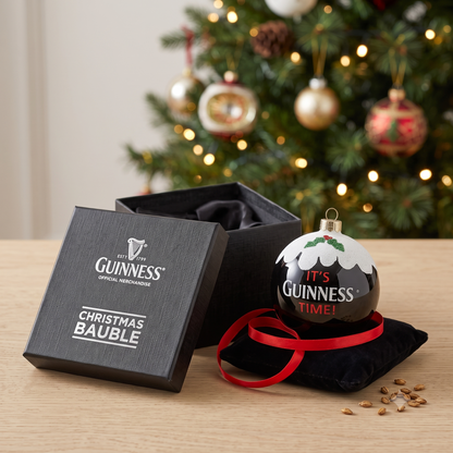 A Guinness Christmas Holiday Pudding Bauble and its box sit on a table with a decorated Christmas tree in the background, creating the perfect festive scene for any tree decoration enthusiast.