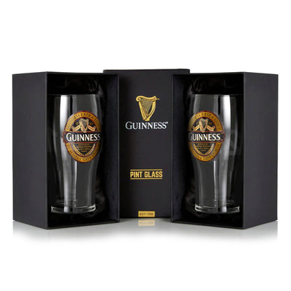 Displayed are two pint glasses in black boxes with gold logos, from the Guinness Classic Pint Glass Twin Pack. The stunning Guinness Classic Ruby Red design adds elegance, making it perfect for enjoying your favorite Extra Stout.