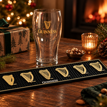 A Guinness From the Archives Gift Set featuring a vintage pint glass behind a Guinness bar mat with six gold-colored harp emblems on a wooden table with holiday decorations.