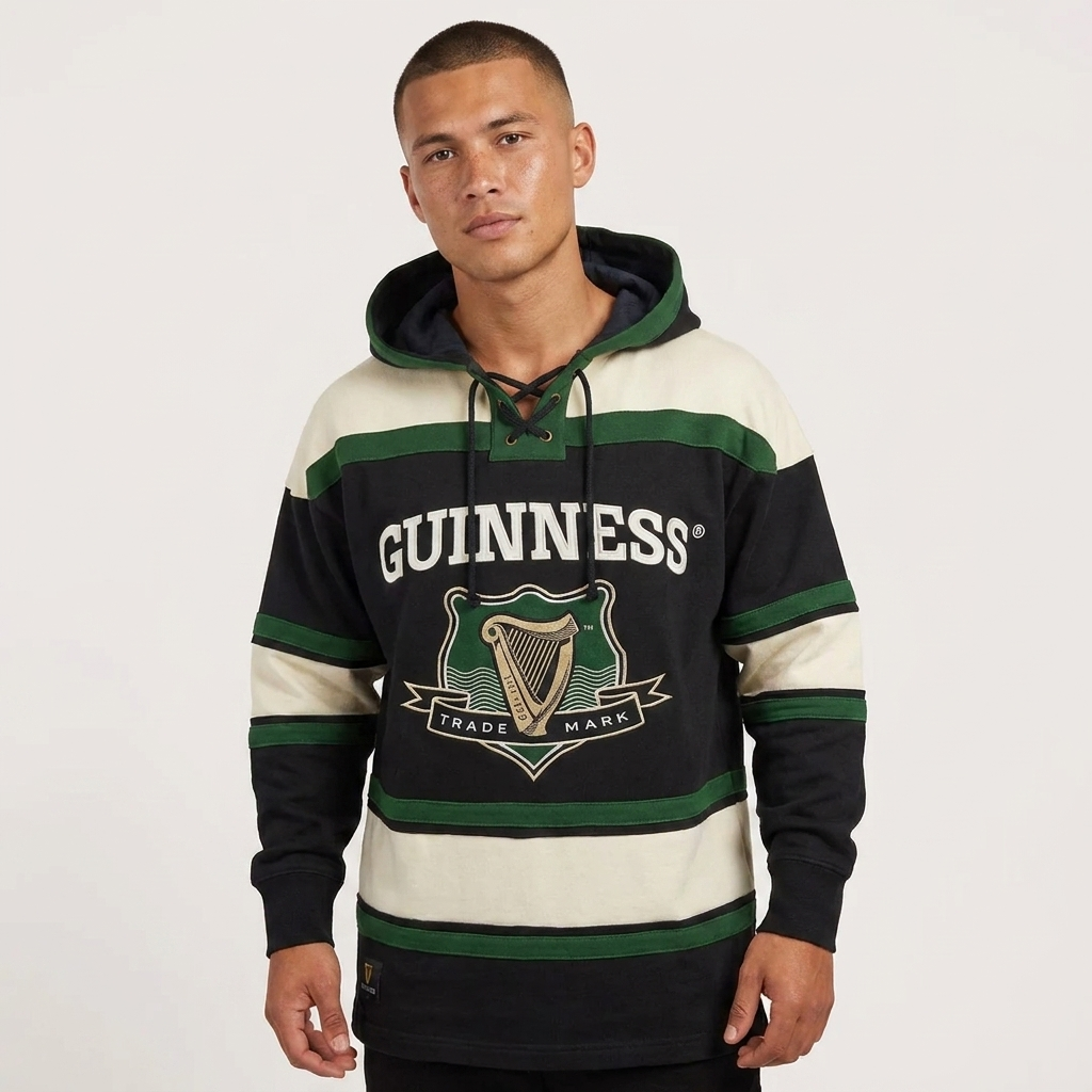 A person stands against a plain background wearing the Guinness Green Hockey Style Hooded Sweatshirt by Guinness, which features black, green, and cream stripes, a harp logo, and the words “Guinness Trade Mark” on the front.