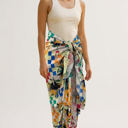 A person wearing a sleeveless beige top, with the Guinness Hawaiian Print Sarong from Guinness Webstore US—a colorful, checkered-patterned scarf—tied around the waist and styled as a vibrant sarong.