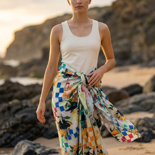 Wearing the Guinness Hawaiian Print Sarong from Guinness Webstore US, a person in a white tank top stands on a rocky beach with cliffs in the background.