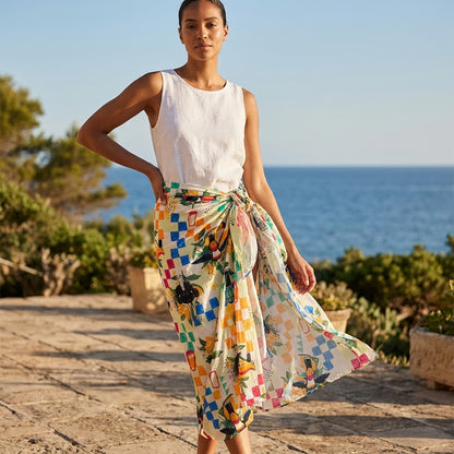 A woman stands outdoors by the sea wearing a white sleeveless top and the Guinness Hawaiian Print Sarong from Guinness Webstore US—a vibrant, floral and geometric patterned wrap skirt, perfect as a chic beach cover-up.