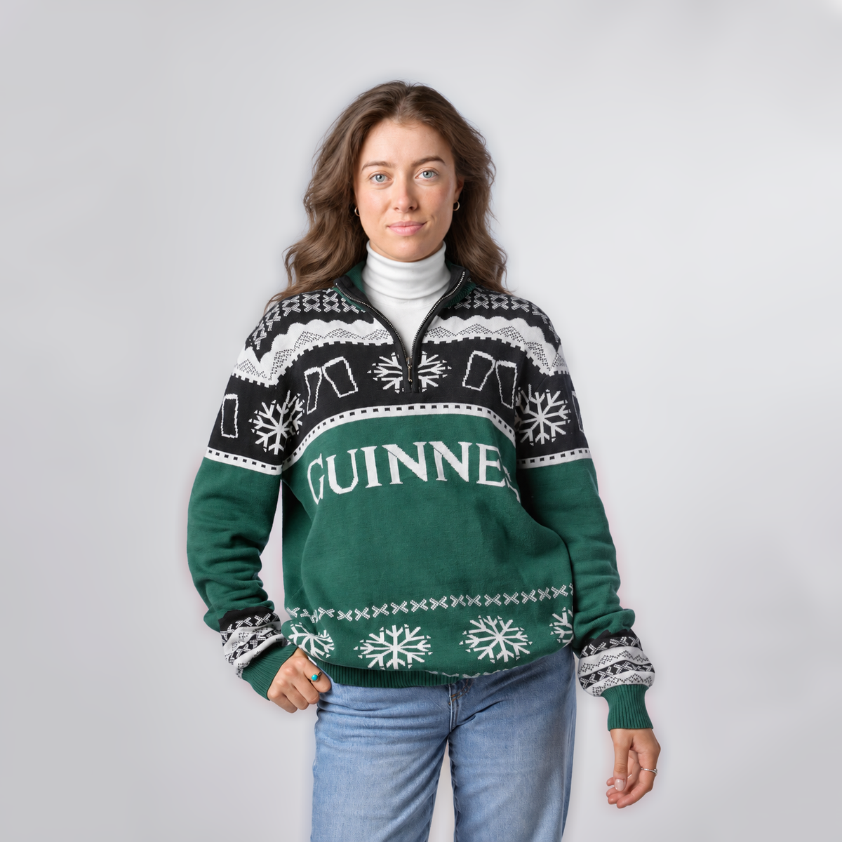 A woman wearing the Guinness Holiday Quarter Zip Sweater in green and black, featuring classic snowflake patterns from Guinness, stands against a plain light background.