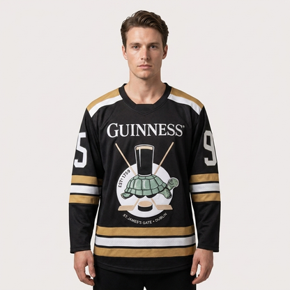 Wearing the Guinness Tortoise Hockey Jersey by Guinness Webstore US—an oversized piece crafted from recycled polyester, featuring the number 95, a turtle graphic, and crossed hockey sticks—a man stands against a plain background.