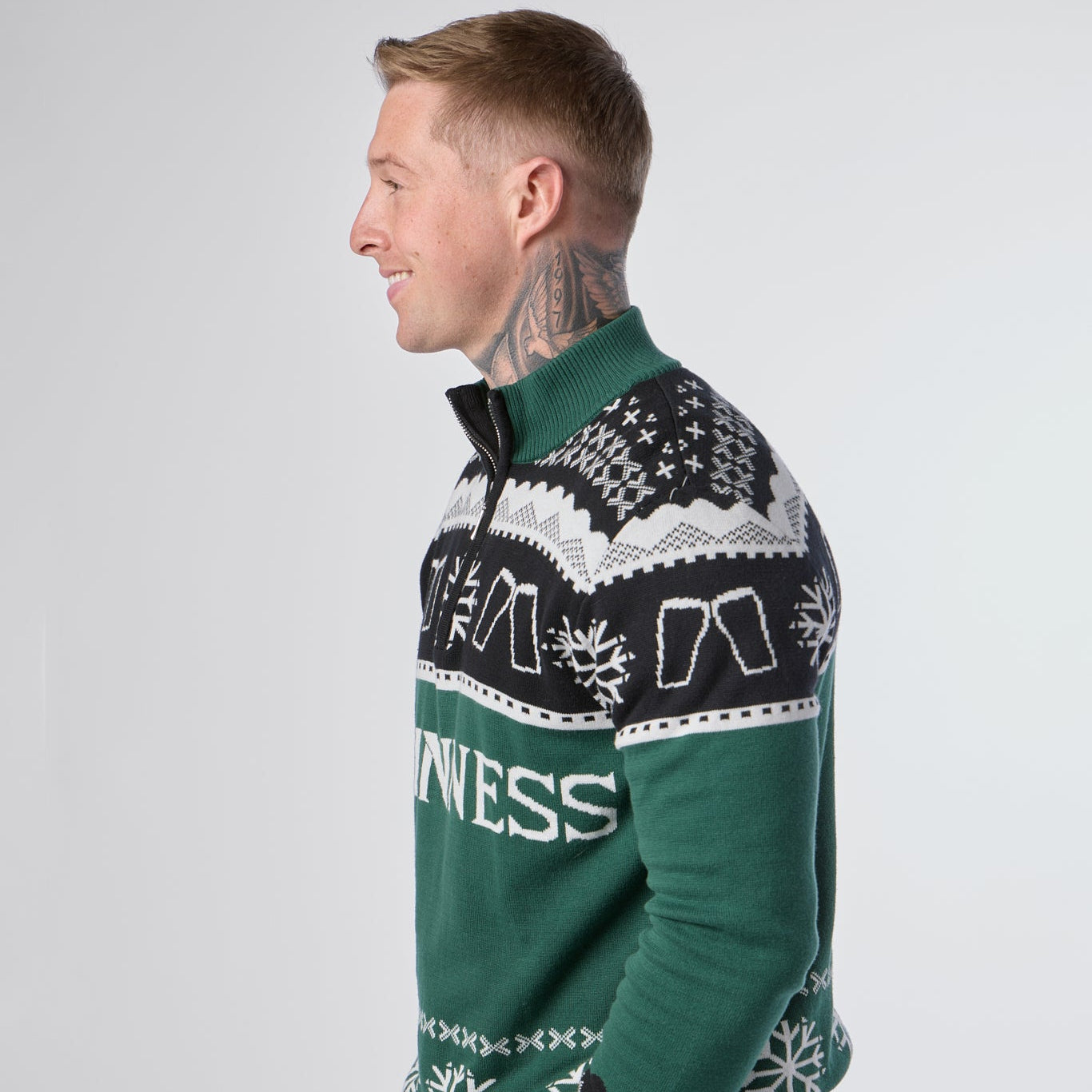 A man with neck tattoos is standing sideways and smiling against a plain background while wearing the Guinness Holiday Quarter Zip Sweater in green, featuring black and white patterns and festive text from the Guinness brand.