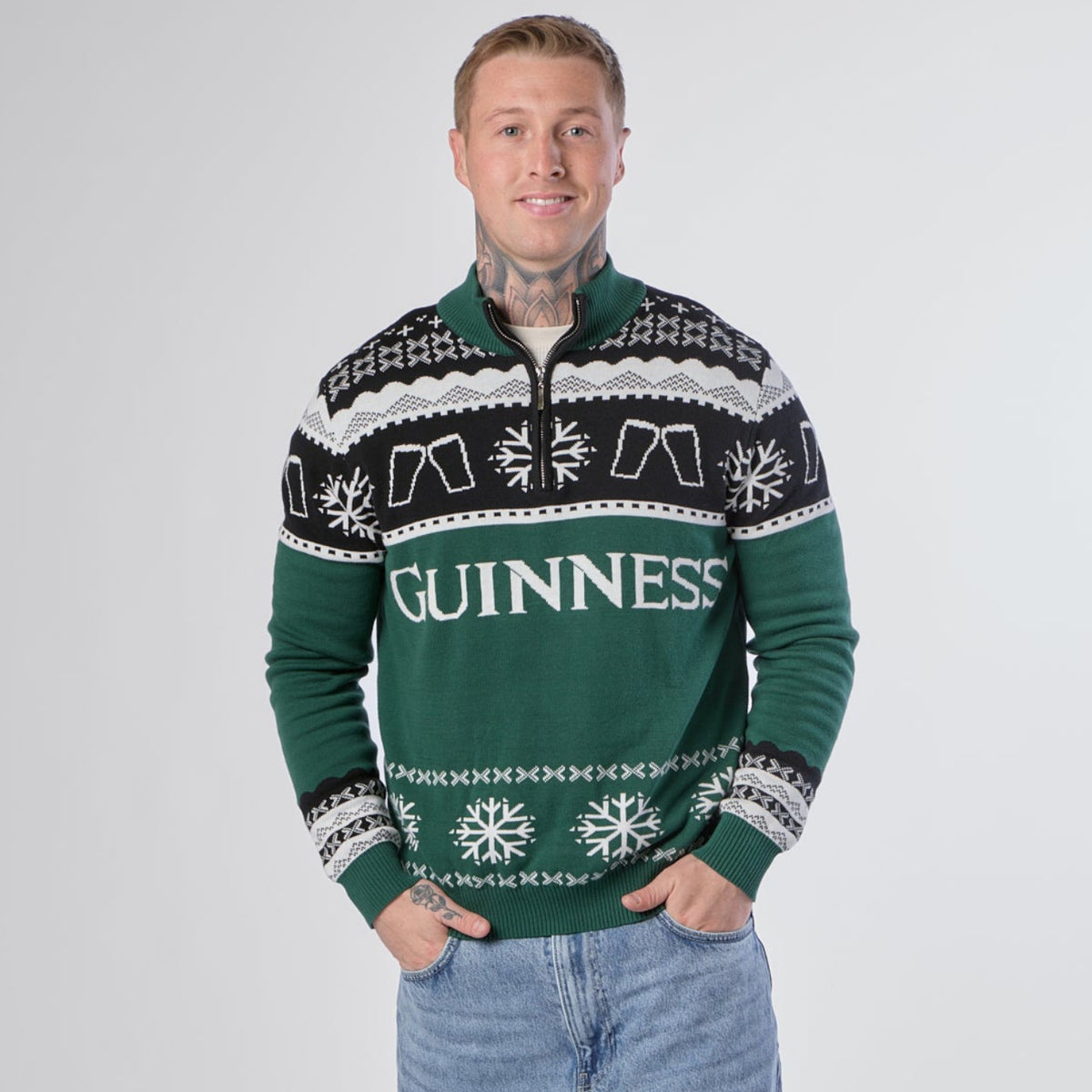 A person stands smiling against a plain white background, wearing the Guinness Holiday Quarter Zip Sweater in green—an iconic Irish Christmas jumper from Guinness featuring black and white patterns—paired with light blue jeans.