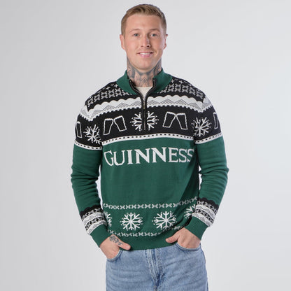 A person stands smiling against a plain white background, wearing the Guinness Holiday Quarter Zip Sweater in green—an iconic Irish Christmas jumper from Guinness featuring black and white patterns—paired with light blue jeans.