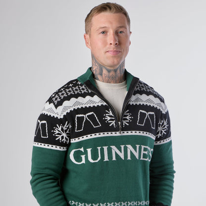 A person with neck tattoos wears the Guinness Holiday Quarter Zip Sweater in green, featuring snowflake patterns and the word “Guinness” on the front, standing against a plain light background.