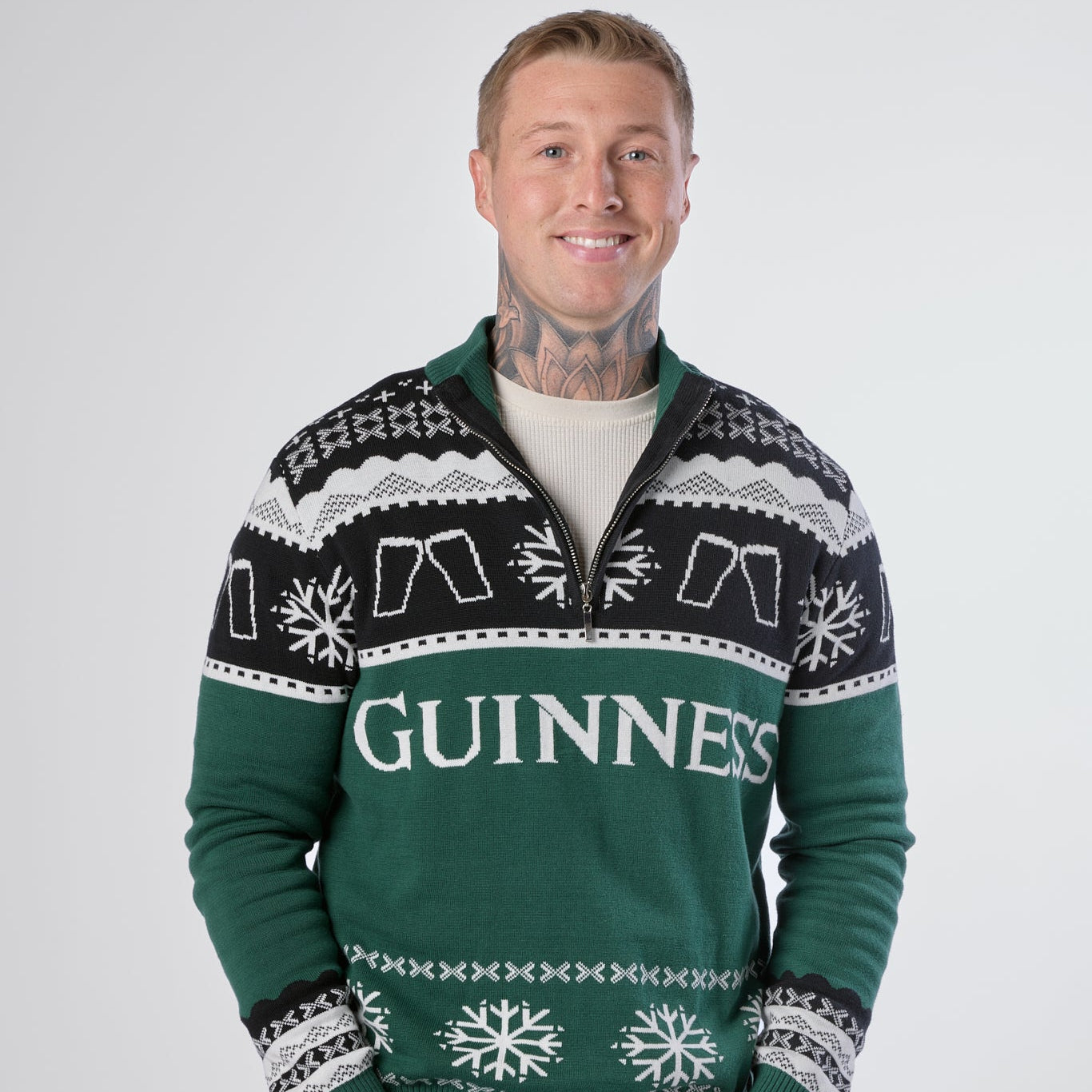 A man wearing the Guinness Holiday Quarter Zip Sweater in green—featuring black and white snowflake patterns from the Guinness brand—stands against a plain light background, smiling at the camera.