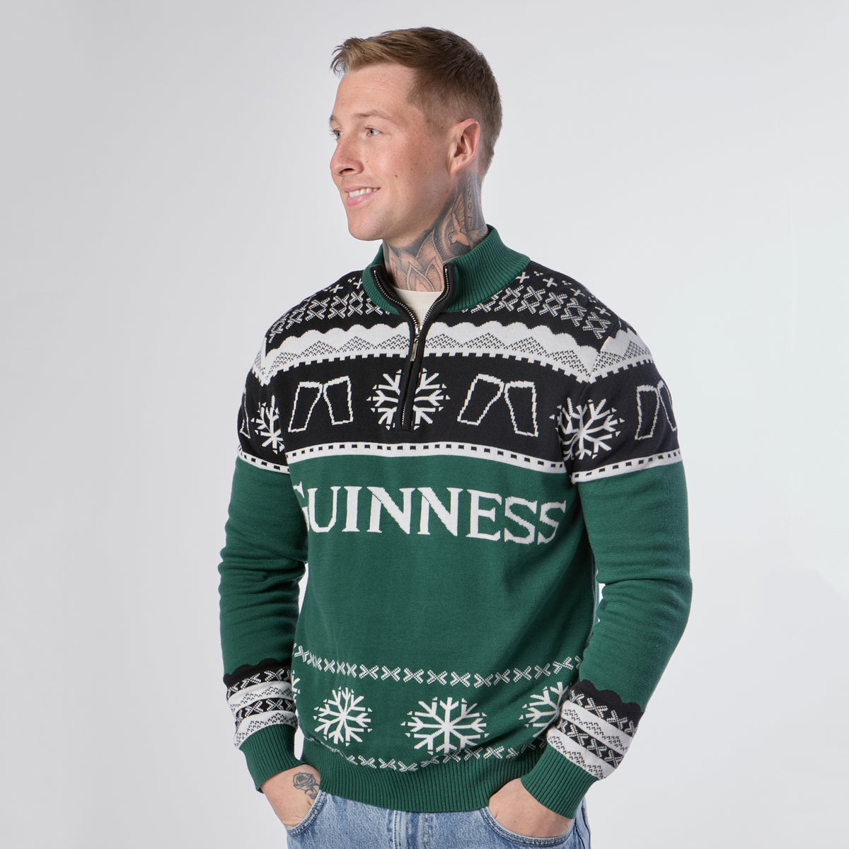 A person stands smiling, wearing the Guinness Holiday Quarter Zip Sweater in green and black—festive knitwear from Guinness featuring “GUINNESS” text, snowflakes, and pint glass graphics—set against a plain light background.