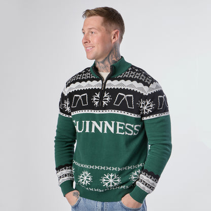 A person stands smiling, wearing the Guinness Holiday Quarter Zip Sweater in green and black—festive knitwear from Guinness featuring “GUINNESS” text, snowflakes, and pint glass graphics—set against a plain light background.