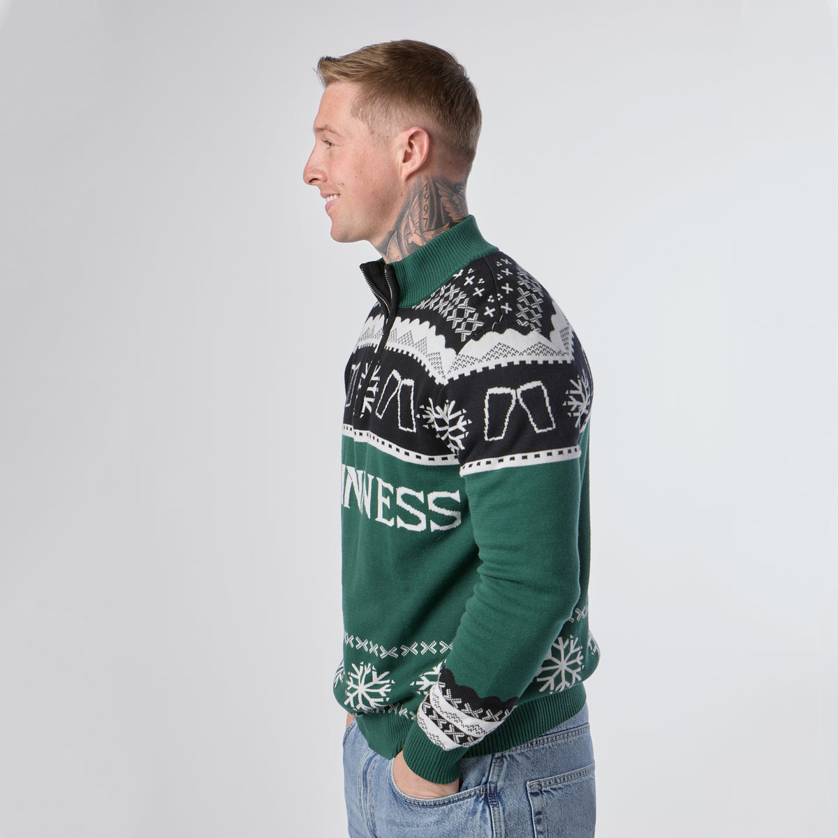 A tattooed man stands in profile with his hands in his pockets, wearing the Guinness Holiday Quarter Zip Sweater—Green & Black by Guinness, featuring festive snowflake patterns and the “Guinness” logo, set against a plain light background.