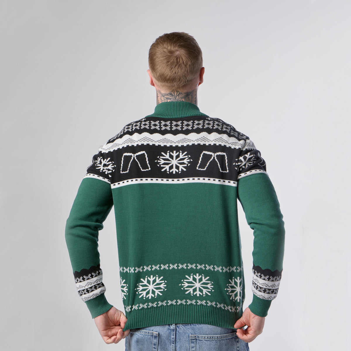 A person stands facing away, wearing the Guinness Holiday Quarter Zip Sweater in green and black with white and black snowflake patterns, paired with light blue jeans, against a plain light background.