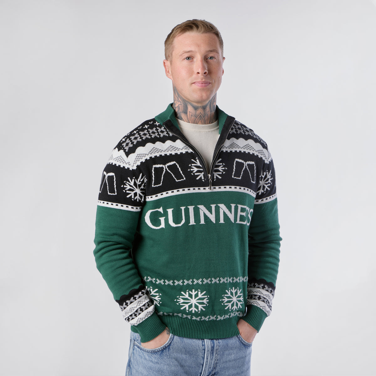 A person wearing the Guinness Holiday Quarter Zip Sweater in green and black, featuring festive snowflake patterns, stands with hands in their jeans pockets against a plain background, stylishly showcasing this seasonal knitwear from Guinness.