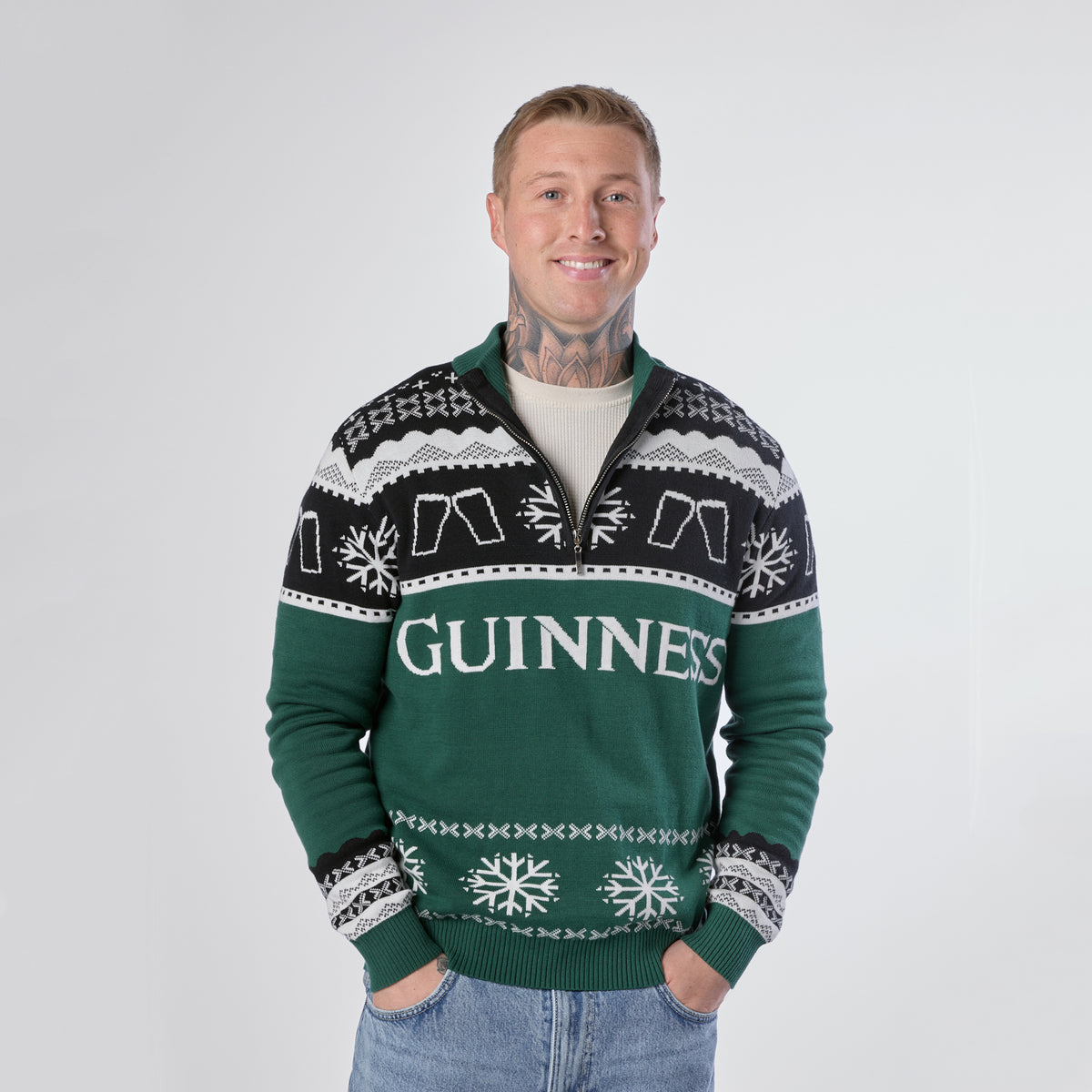 A person stands against a plain backdrop wearing the Guinness Holiday Quarter Zip Sweater in green and black, featuring festive snowflake patterns and the word “Guinness” across the front, with their hands clasped in front.