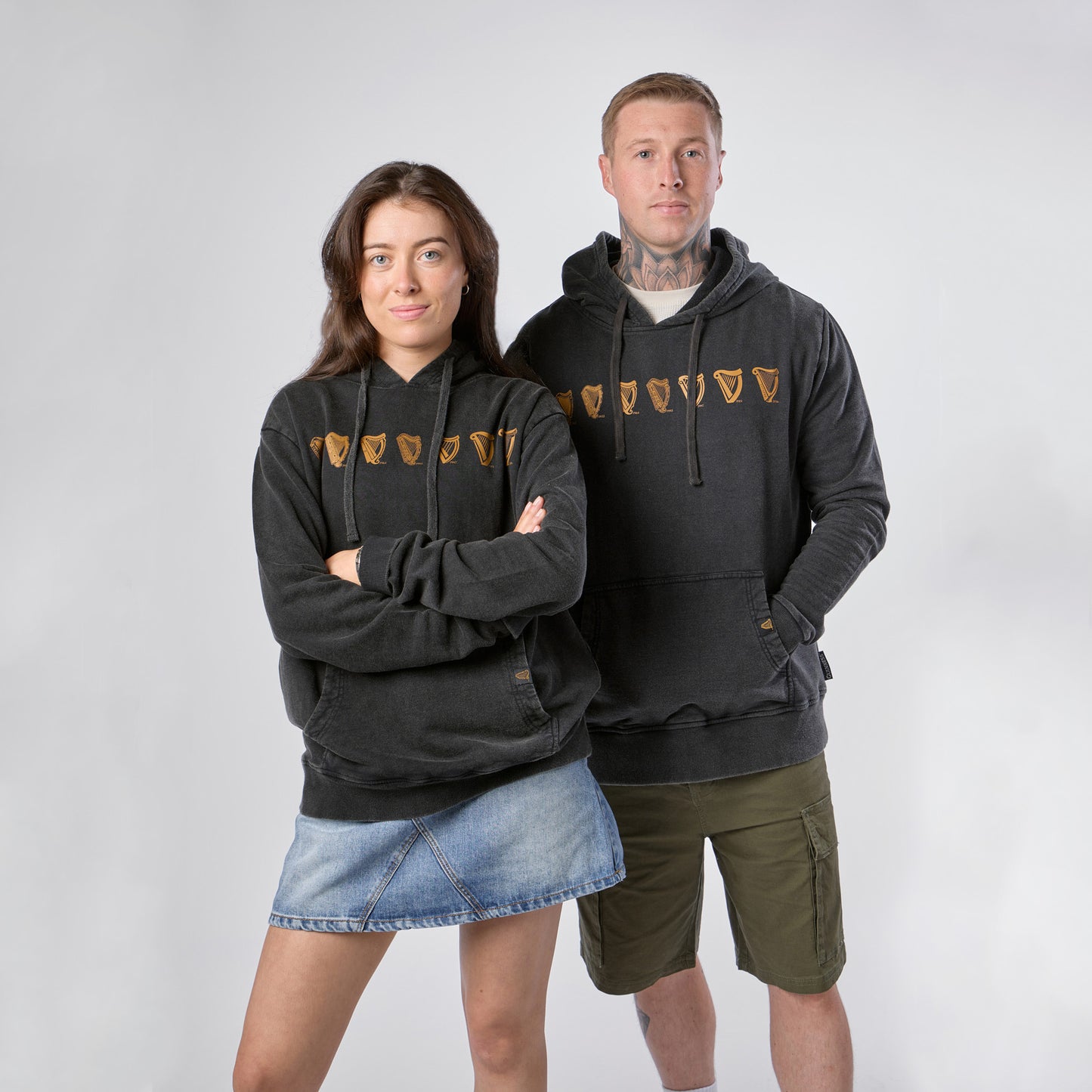 Two people stand side by side against a plain background, each showcasing the Guinness Evolution of the Harp Distressed Hoodie – Black from Guinness Webstore US with its distinctive gold shield design; one styles the hoodie with a denim skirt, while the other pairs it with khaki shorts.
