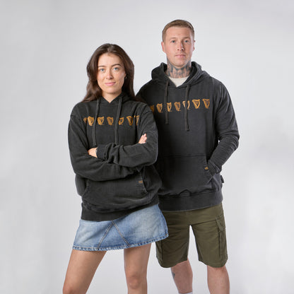 Two people stand side by side against a plain background, each showcasing the Guinness Evolution of the Harp Distressed Hoodie – Black from Guinness Webstore US with its distinctive gold shield design; one styles the hoodie with a denim skirt, while the other pairs it with khaki shorts.