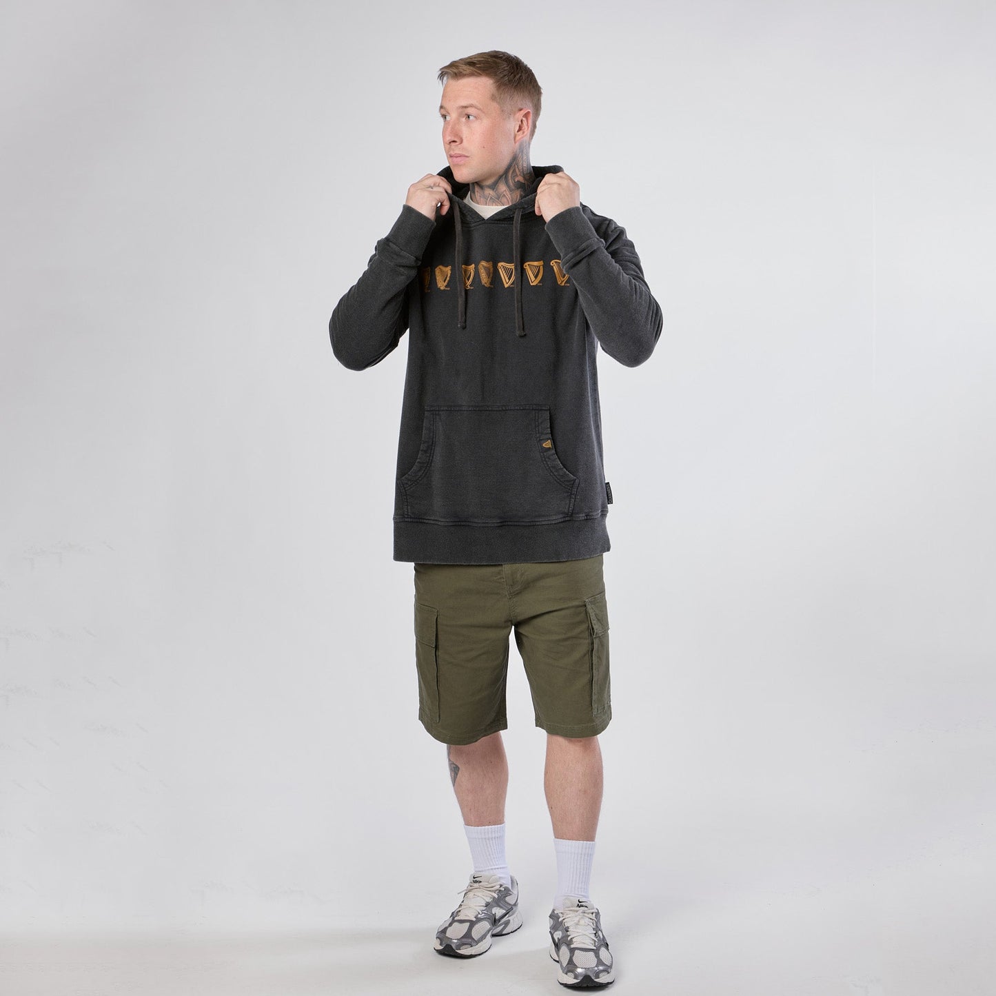 A person wearing a Guinness Evolution of the Harp Distressed Hoodie – Black from Guinness Webstore US, paired with olive green cargo shorts, white socks, and sneakers, stands against a plain white background while holding the hoodie’s drawstrings.