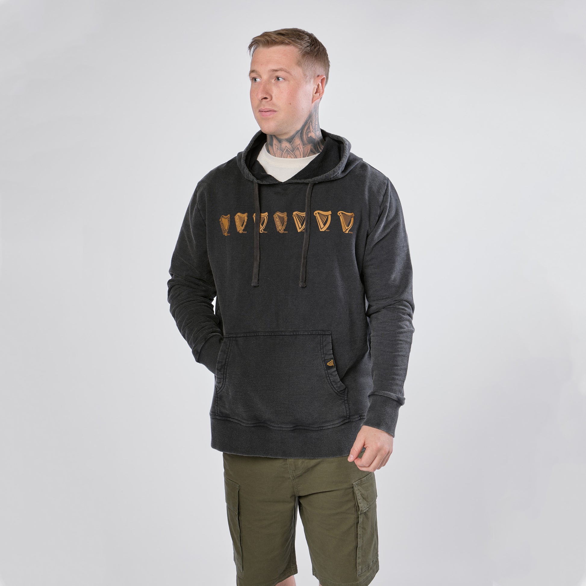 A man wearing the Guinness Evolution of the Harp Distressed Hoodie in black from Guinness Webstore US and olive green shorts stands against a plain light background, looking slightly to the side.