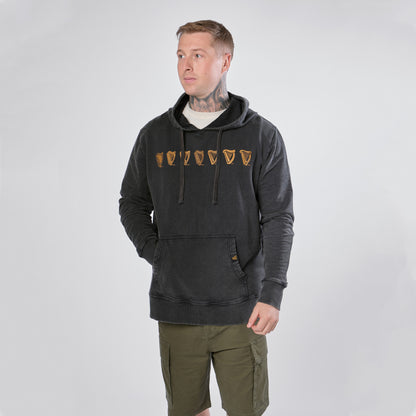 A man wearing the Guinness Evolution of the Harp Distressed Hoodie in black from Guinness Webstore US and olive green shorts stands against a plain light background, looking slightly to the side.