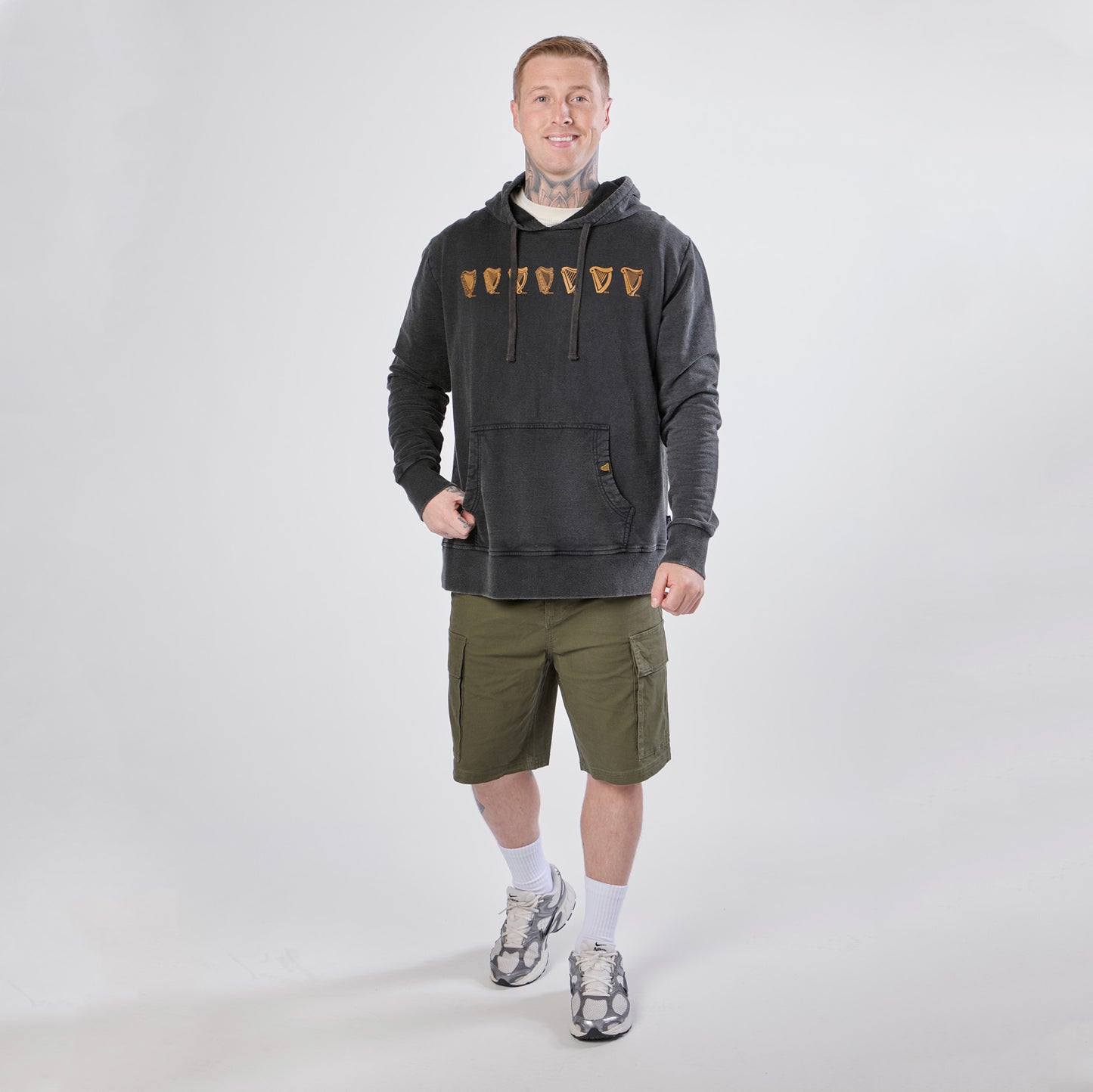 A man stands against a plain background wearing a black Guinness Evolution of the Harp Distressed Hoodie from Guinness Webstore US with graphic designs, green cargo shorts, white socks, and gray sneakers.