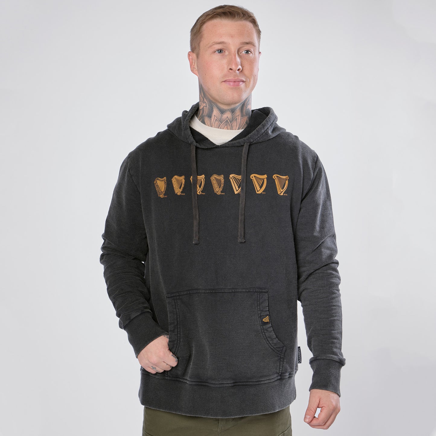 A man wearing a black Guinness Evolution of the Harp Distressed Hoodie from Guinness Webstore US, featuring gold pint glass graphics, stands against a plain light background with one hand in his pocket.