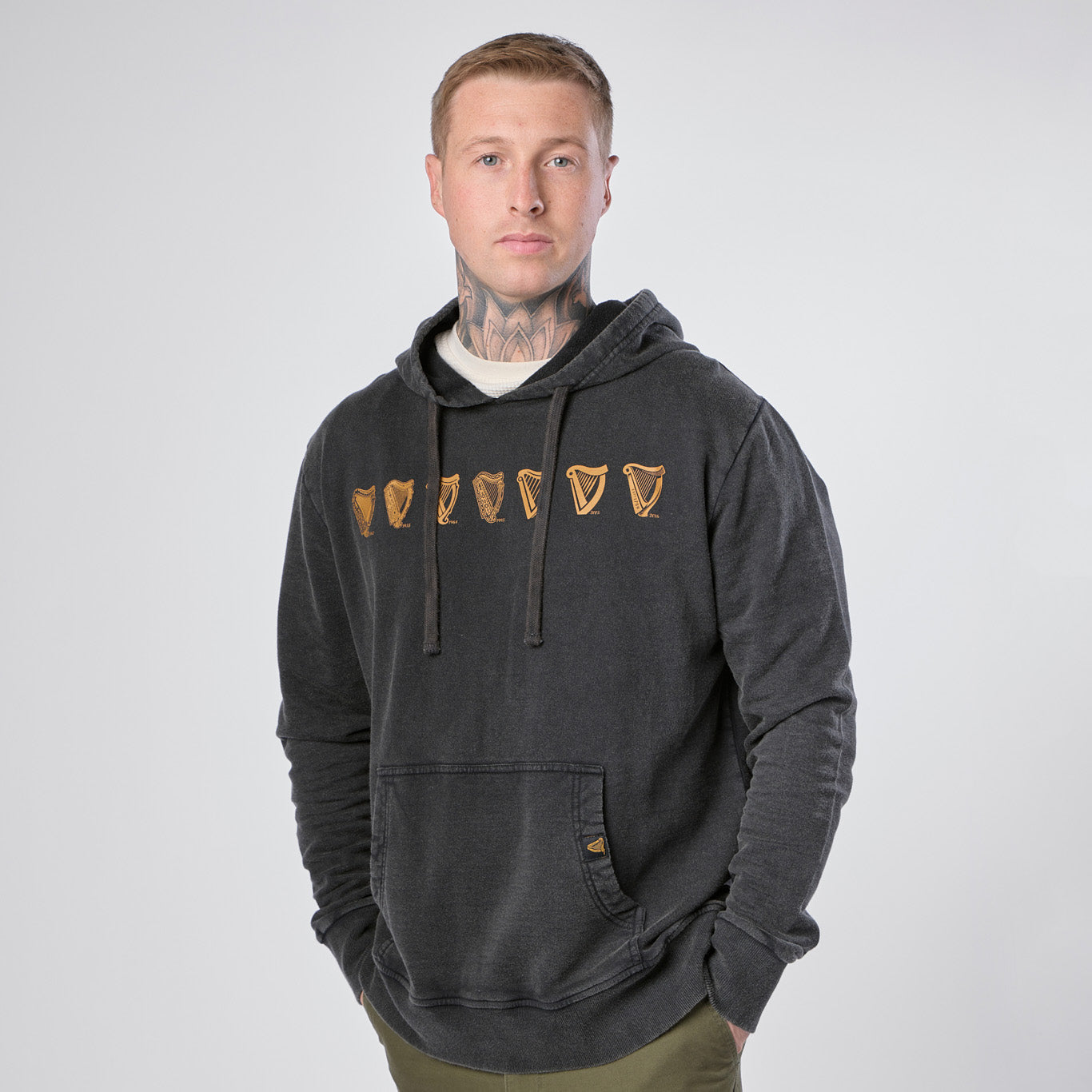 A man wearing the Guinness Webstore US “Guinness Evolution of the Harp Distressed Hoodie” in black stands against a plain white background, hands in pockets, looking directly at the camera.