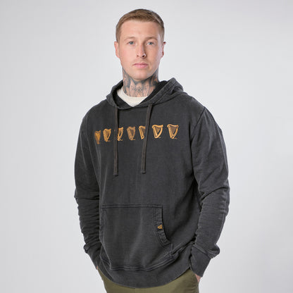 A man wearing the Guinness Webstore US “Guinness Evolution of the Harp Distressed Hoodie” in black stands against a plain white background, hands in pockets, looking directly at the camera.