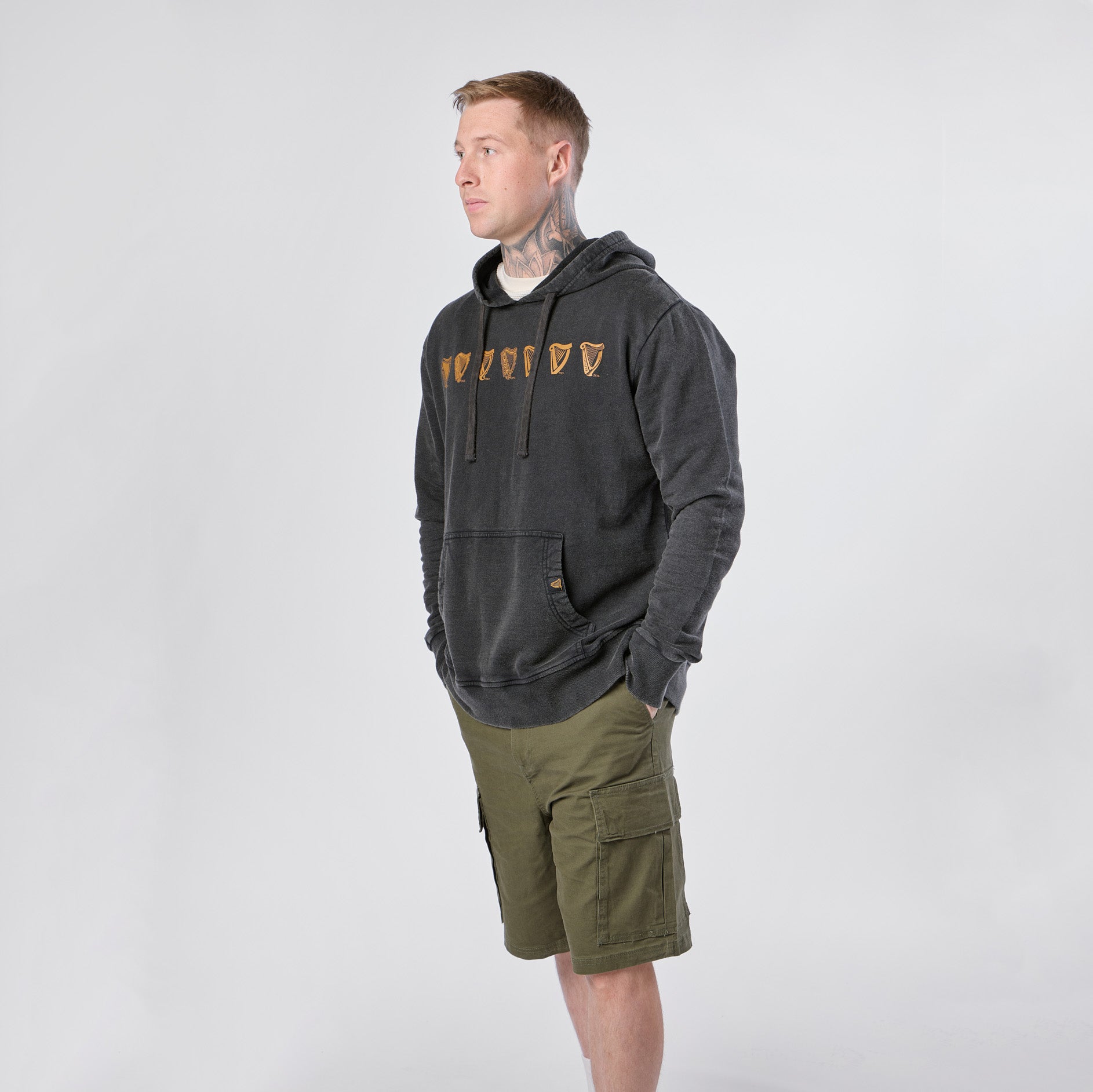 A person wearing the Guinness Evolution of the Harp Distressed Hoodie in black from Guinness Webstore US and green cargo shorts stands against a plain white background.