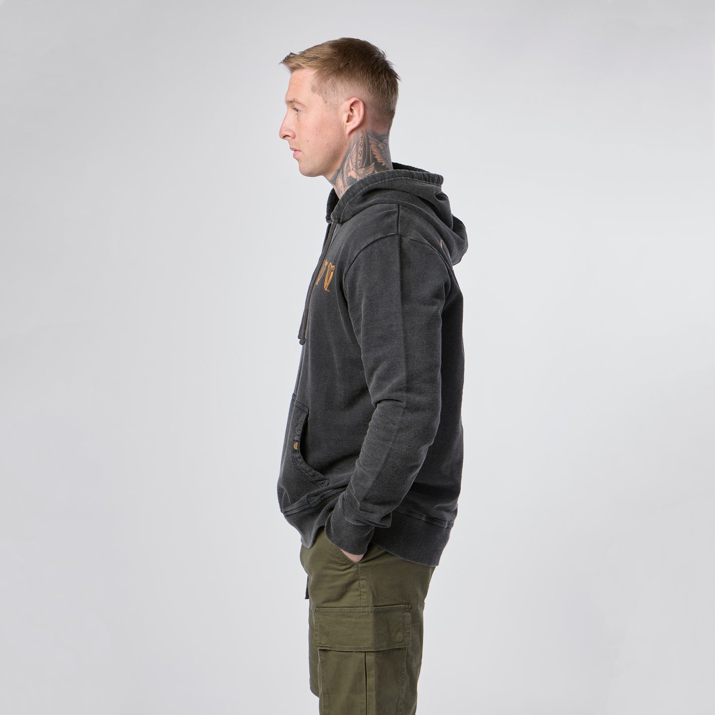 A man with short hair and neck tattoos stands in profile, wearing the Guinness Evolution of the Harp Distressed Hoodie in black from Guinness Webstore US and green cargo pants, his hands in his pockets against a plain light background.
