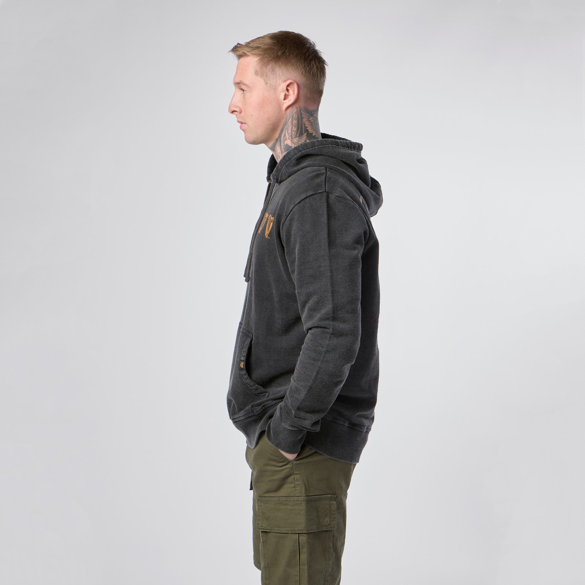 A man with short hair and neck tattoos stands in profile, wearing the Guinness Evolution of the Harp Distressed Hoodie in black from Guinness Webstore US and green cargo pants, his hands in his pockets against a plain light background.