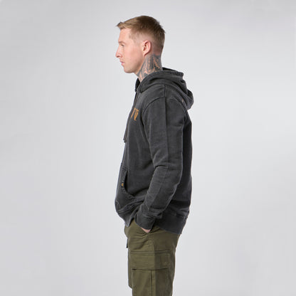 A man with short hair and neck tattoos stands in profile, wearing the Guinness Evolution of the Harp Distressed Hoodie in black from Guinness Webstore US and green cargo pants, his hands in his pockets against a plain light background.