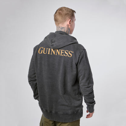A person stands facing away, wearing the Guinness Evolution of the Harp Distressed Hoodie – Black from Guinness Webstore US, which features “GUINNESS” in gold lettering on the back. The background is plain and light-colored.