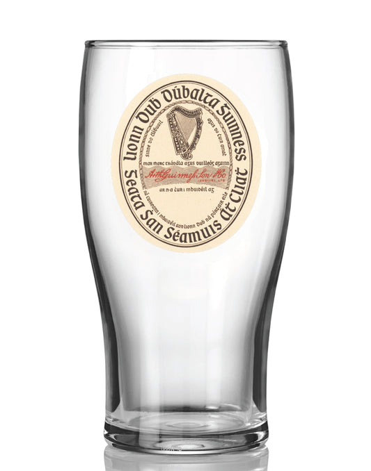 The Guinness Vintage Label Pint Glass from Guinness Webstore US is a clear tulip pint glass displaying a round, vintage-style Guinness Extra Stout label with the iconic harp logo and bilingual English-Irish text.
