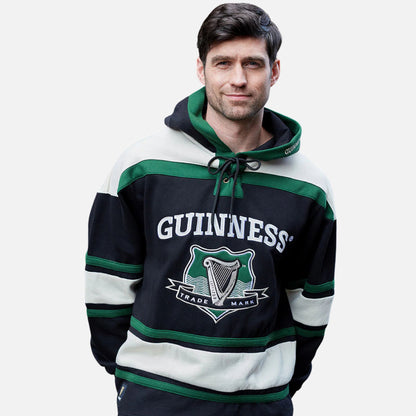 A man wears the Guinness Green Hockey Style Hooded Sweatshirt featuring "GUINNESS" and a harp logo on the front, crafted from a heavy-weight cotton blend, standing against a plain light background.