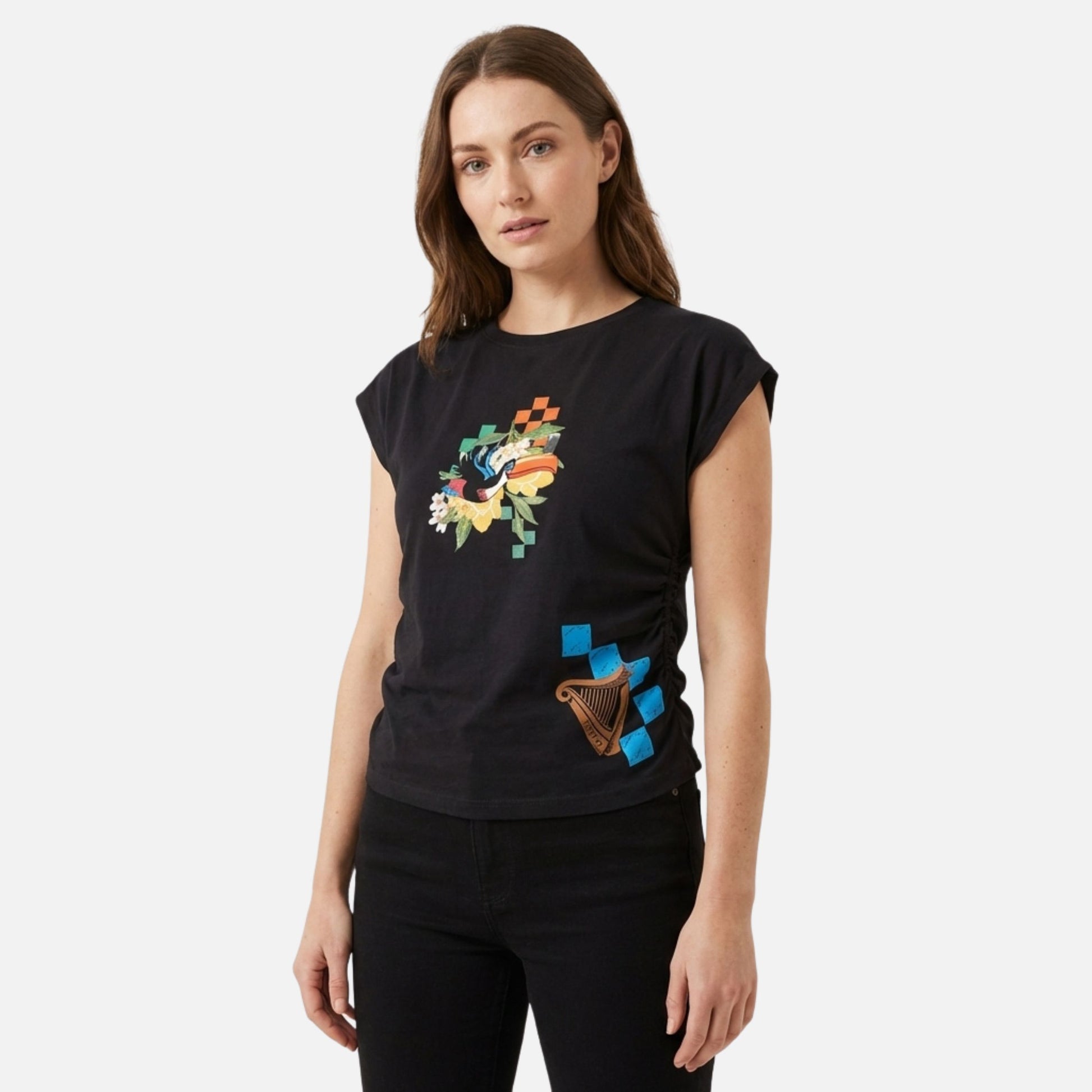 A woman stands against a plain background wearing the Guinness Toucan Ruched Ladies Black Tee from Guinness Webstore US, featuring a vibrant toucan graphic and colorful abstract designs, paired with black pants.