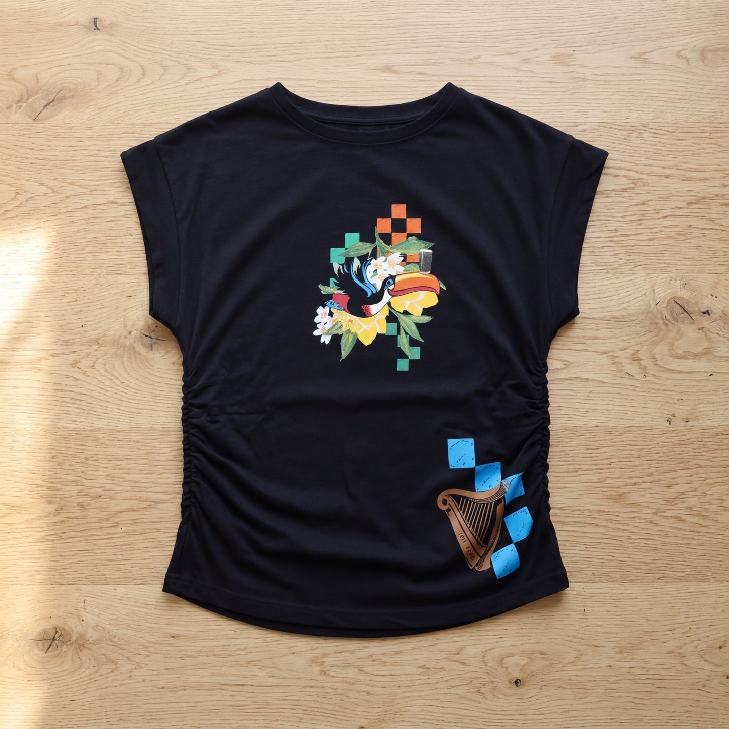 The Guinness Toucan Ruched Ladies Black Tee from Guinness Webstore US features colorful abstract floral and geometric chest designs, plus a brown harp with blue squares near the bottom right. The shirt is displayed on a wooden surface.