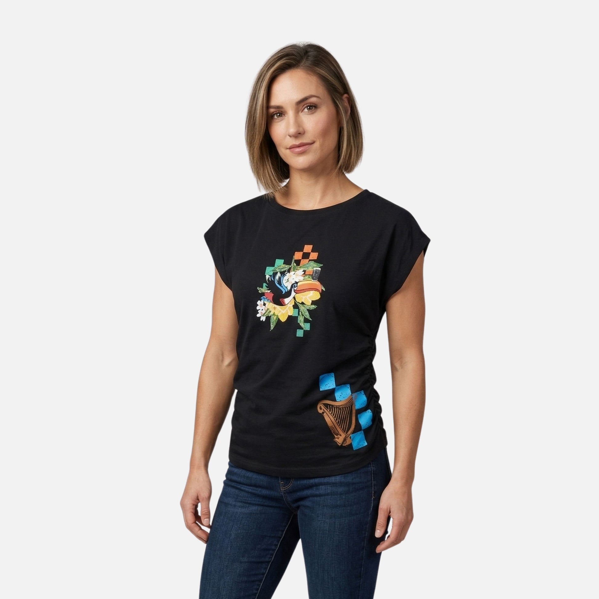 A woman stands against a plain background, wearing the Guinness Toucan Ruched Ladies Black Tee from Guinness Webstore US and dark blue jeans.