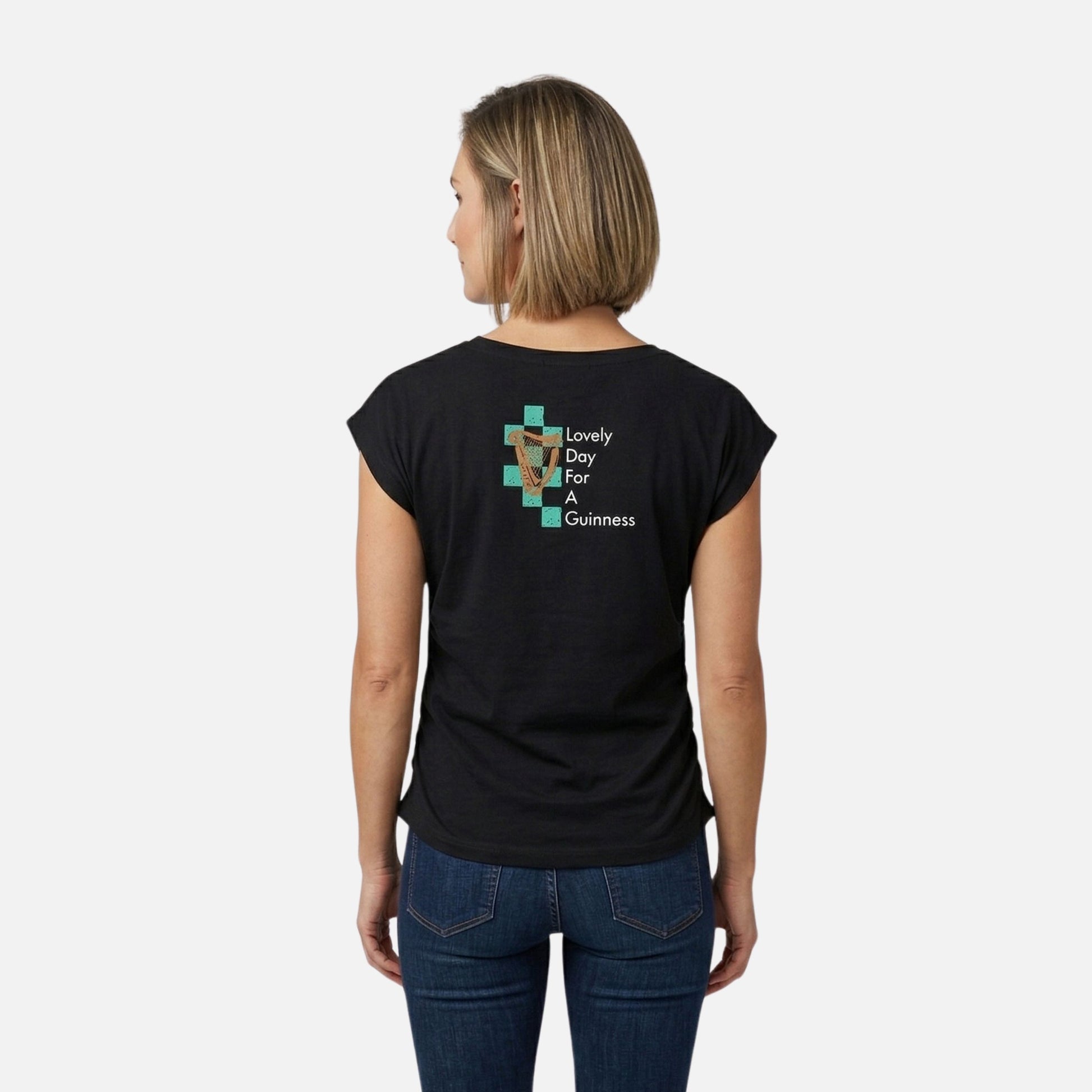A woman faces away, wearing the Guinness Toucan Ruched Ladies Black Tee from Guinness Webstore US, which features a toucan graphic and "Lovely Day For A Guinness" text on the back.