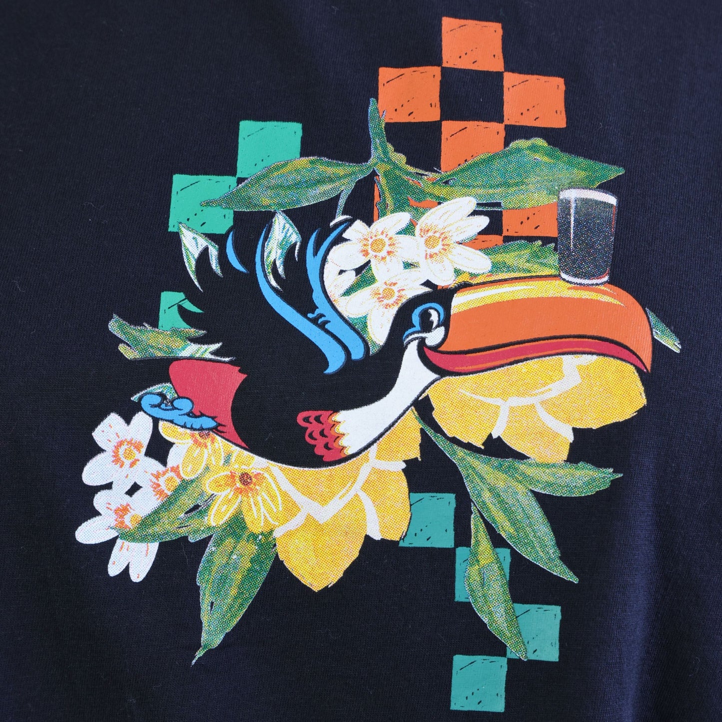 The Guinness Toucan Ruched Ladies Black Tee from Guinness Webstore US features a vibrant toucan graphic with colorful geometric shapes and white-yellow flowers on a dark background, inspired by the classic Guinness aesthetic.