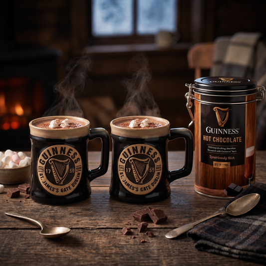 Two mugs of Guinness Hot Chocolate from the Guinness Night In Gift Set, topped with marshmallows, next to a canister of mix, chocolate chunks, and two spoons on a wooden table.