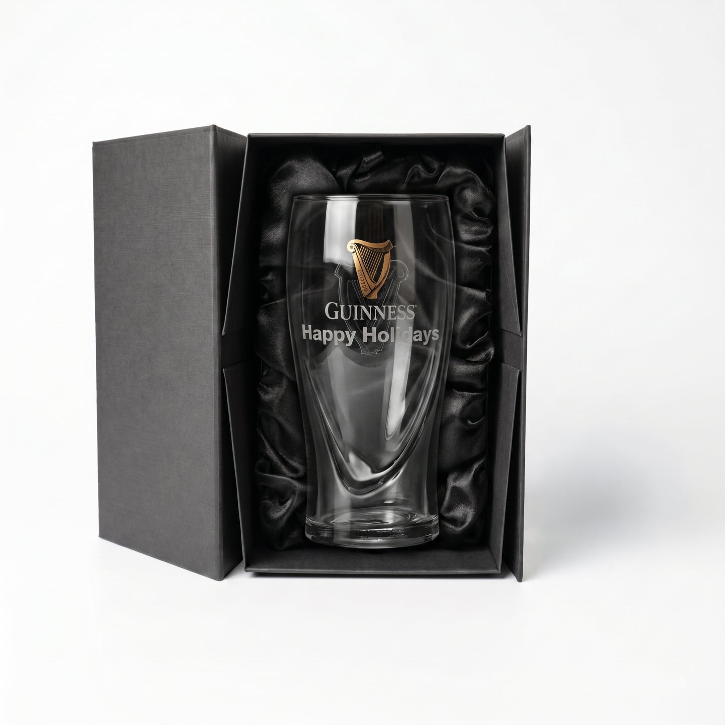 The Guinness Pint Glass Luxury Gift Box from Guinness Webstore US features a boxed pint glass adorned with a gold harp logo and “Happy Holidays” text, elegantly presented in a premium gift box lined with black satin, displayed against a white background.