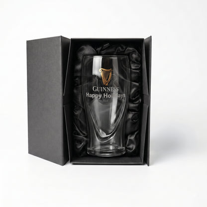 The Guinness Pint Glass Luxury Gift Box from Guinness Webstore US features a boxed pint glass adorned with a gold harp logo and “Happy Holidays” text, elegantly presented in a premium gift box lined with black satin, displayed against a white background.