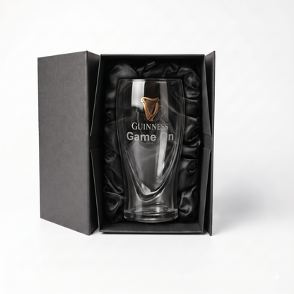 Showcased in an open luxury gift box lined with black satin, the Guinness Webstore US Guinness Pint Glass Luxury Gift Box features a clear pint glass adorned with a gold Guinness logo and “Game On” text, making for an impressive presentation.